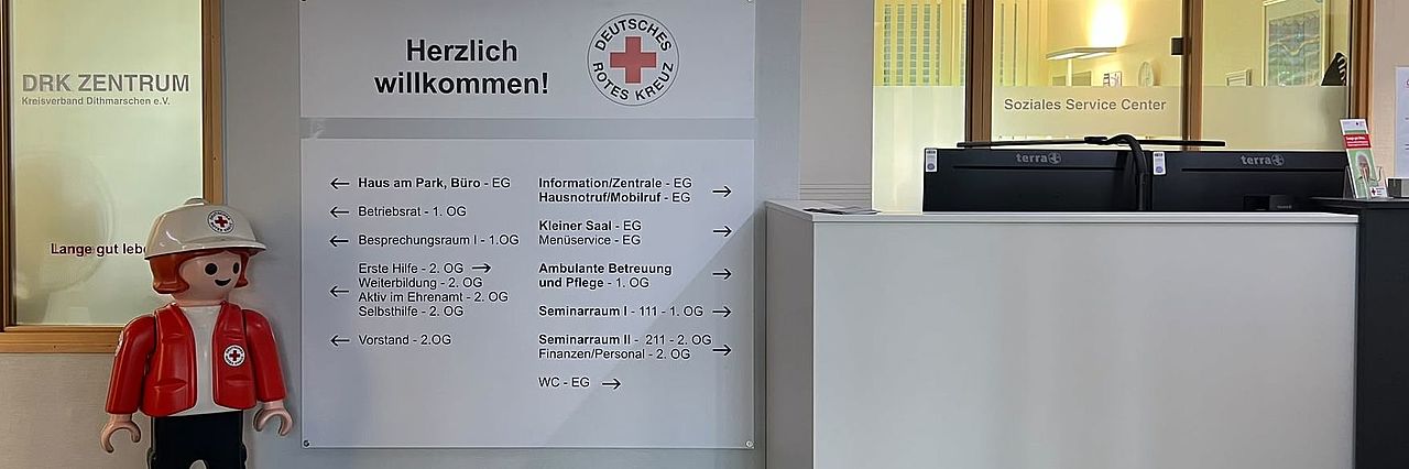 Foyer Kreisverband Blick in den Eingangs-/Tresenbereich. Links Soziales Service Center. Kleine Playmobilfigur, Kind in DRK Kleidung. Helle Übersichtstafel mit Hinweisen zu den Bereichen im Kreisverband. Rechts Teile des Empfangstresen