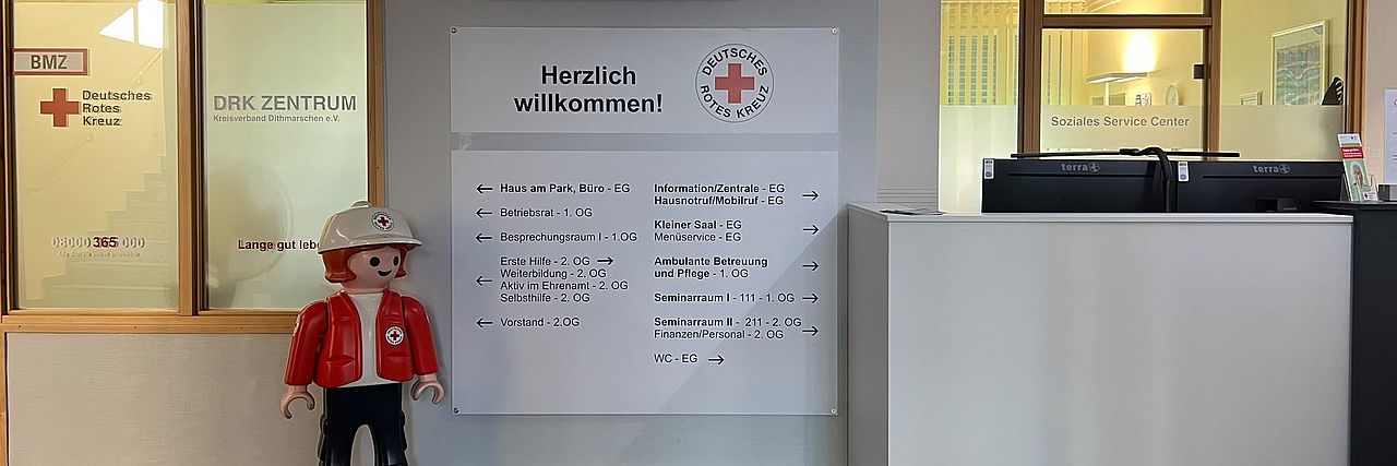 Foyer Kreisverband Blick in den Eingangs-/Tresenbereich. Links Soziales Service Center. Kleine Playmobilfigur, Kind in DRK Kleidung. Helle Übersichtstafel mit Hinweisen zu den Bereichen im Kreisverband. Rechts Teile des Empfangstresen
